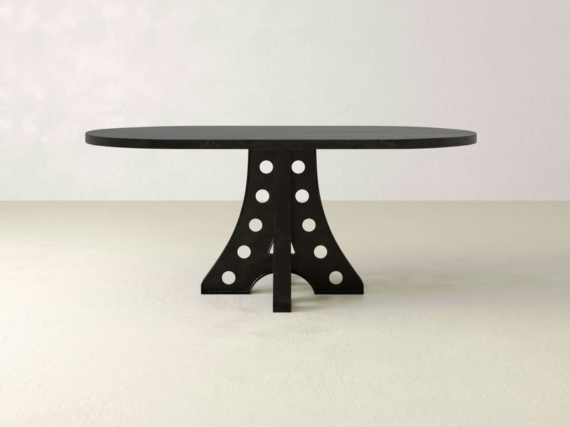 The Amelia Oval Dining Table - Charred Ember by James+James is a modern black table with a unique central support design featuring circular cutouts. It stands on a smooth, light-colored floor against a white wall, and exudes a sleek and contemporary appearance.