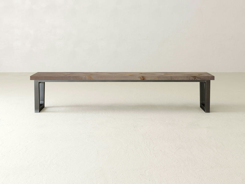 The Trapezoid Bench - Barn Wood by James+James, with its rectangular wooden seat and modern metal legs, sits against a plain, light-colored wall. The minimalist design features a natural wood finish and dark metal supports, creating a sleek and contemporary look.