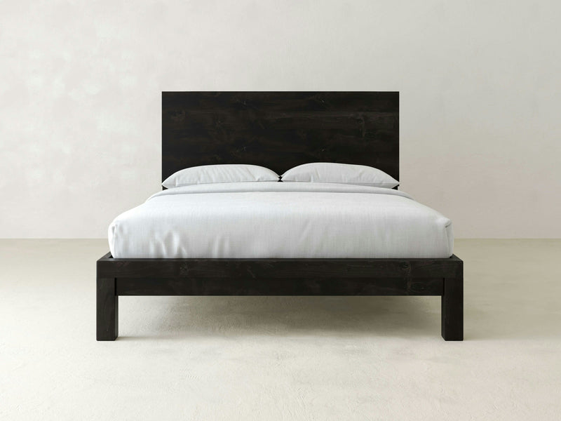 The Sawyer Bed by James+James, a minimalist dark wooden bed frame crafted from solid hardwood with a simple headboard, is placed against a plain light-colored wall. The bed is neatly made with white bed sheets and pillows, accentuating the timeless sophistication and clean design of the room.