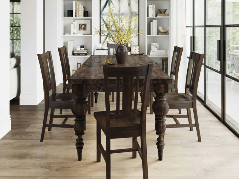 A dining area featuring the Bailey Dining Table - Tobacco by James+James accompanied by six matching chairs, decorated with a vase of yellow flowers. The room boasts light wooden flooring, white walls, and large windows offering an outdoor view. Bookshelves in the background display various items.