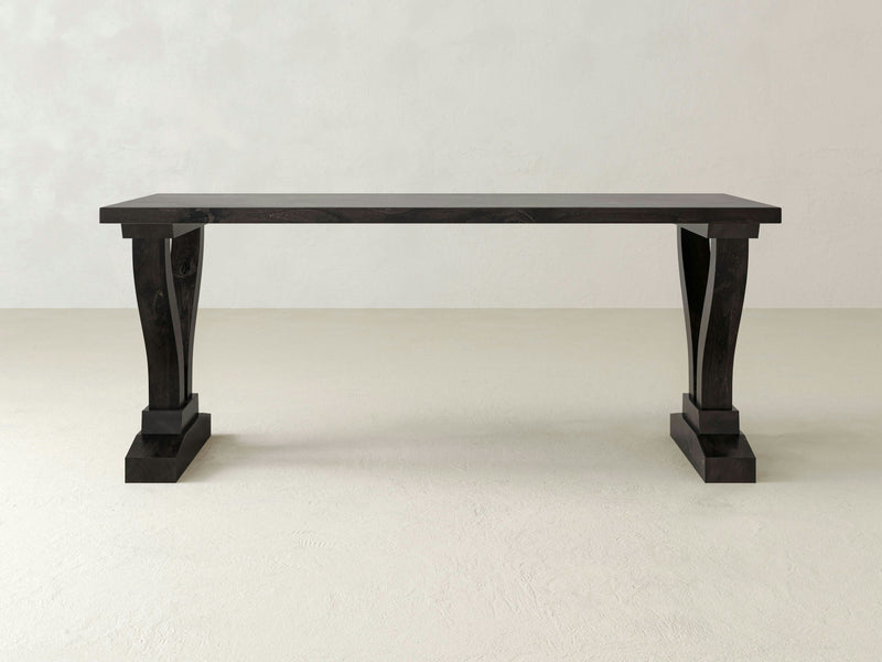 The James+James Vera Desk - Charred Ember is a black, rectangular wooden table with a thick tabletop and two ornate pedestal legs. It is set against a plain off-white background, showcasing its minimalist design that merges modern and classic elements with a sturdy, functional appearance.