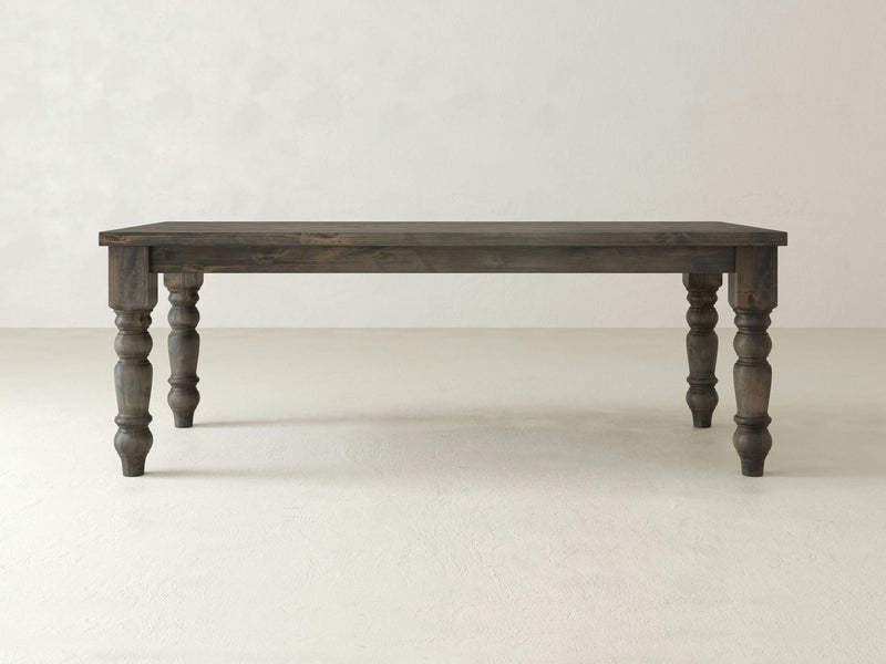 The Baluster Dining Table - Deep Grey by James+James is set against a plain, light-colored background. This rectangular wooden table boasts four intricately carved legs and has a rustic, vintage appearance with its dark, weathered finish.