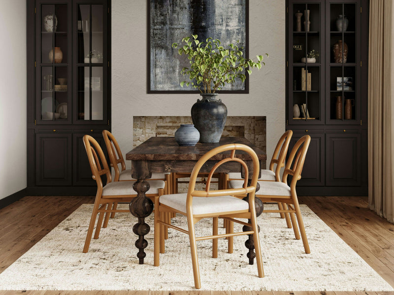 A dining room featuring the Olivia Dining Table in Tobacco by James+James, surrounded by six light wooden chairs on a beige rug. A large plant in a dark vase and a smaller blue vase adorn the table. The backdrop includes two dark cabinets with glass doors and a large abstract painting.