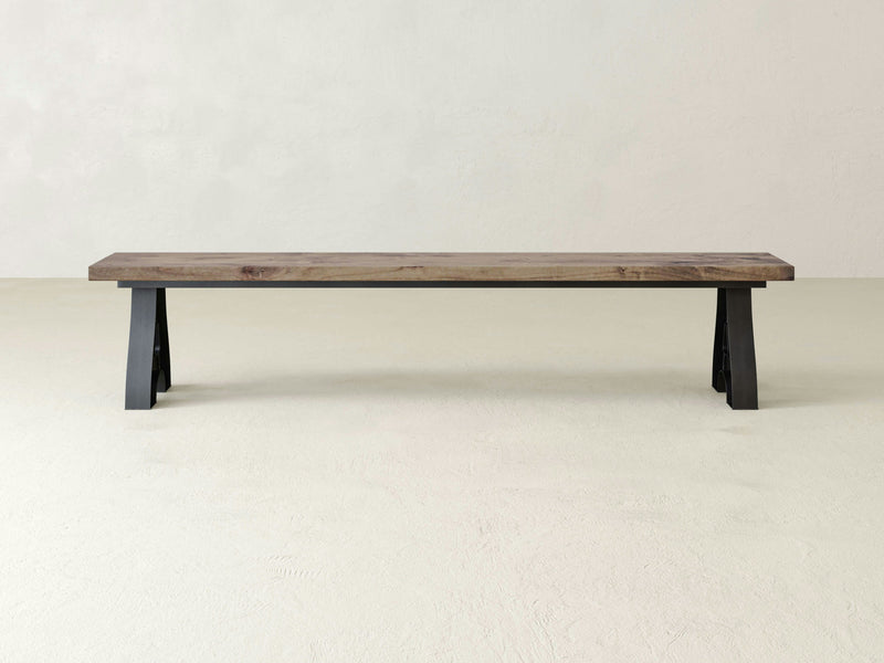 The James+James Wishbone Bench boasts a handcrafted design made from solid hardwood with a natural finish, featuring a flat rectangular seat and durable black metal legs. It sits elegantly on a light-colored floor against a plain off-white background.