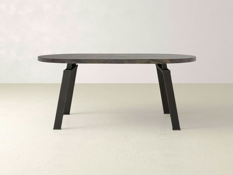 The Fulton Oval Dining Table - Deep Grey by James+James features a minimalist oval top in a deep grey finish and four angled black metal legs, set against a plain, light-colored background. The table's modern design gives it a sleek and contemporary appearance.