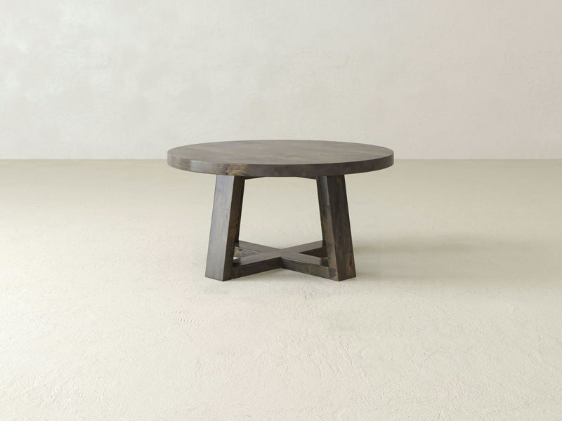 A simple, round Rae Coffee Table - Deep Grey by James+James sits on a plain, light-colored floor. The table has a distinct, sturdy base featuring four angled legs that form an intersecting X shape at the bottom. The background is a plain, off-white wall.