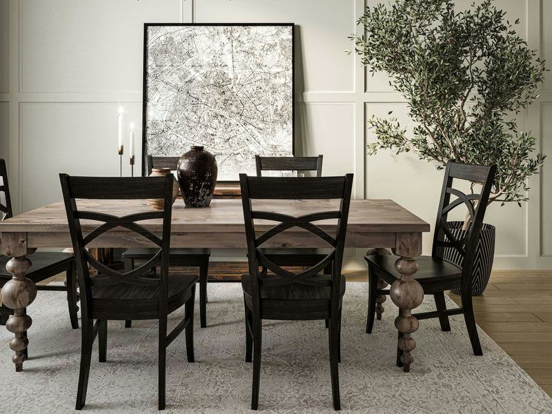 A dining room with the Olivia Expandable Dining Table in Barn Wood by James+James, surrounded by six dark cross-back chairs. On the table are two tall candles and a large ceramic vase. A large abstract art piece leans against the wall, and a leafy indoor plant sits beside the table.