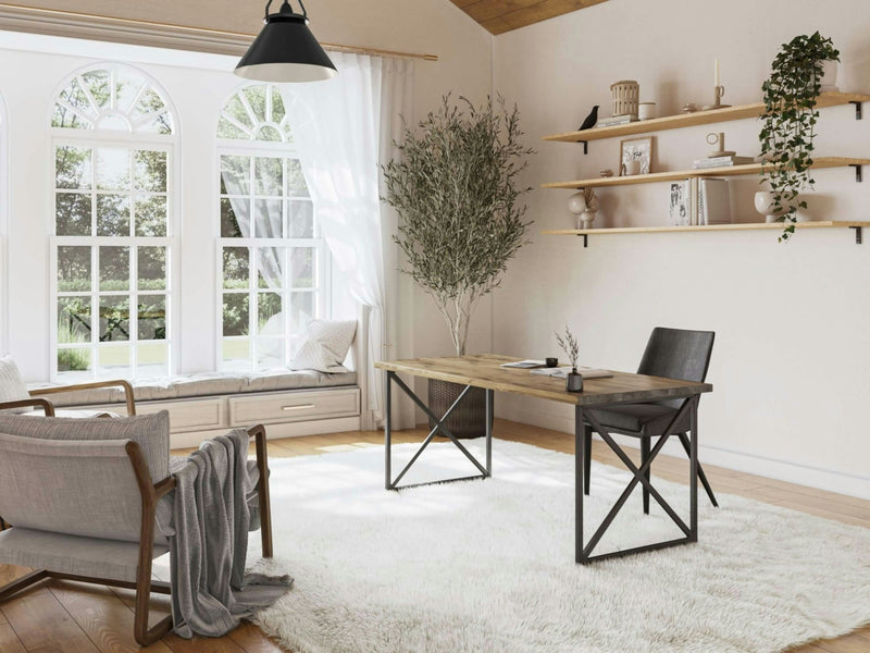 A bright, airy home office features the Luca Desk in Harvest Wheat by James+James, designed with a wooden top and black metal legs. The space also includes a dark gray chair and a beige rug. A large window with arched panes lets in natural light, while shelves adorned with decor and plants, an armchair, and a potted tree complete the space.