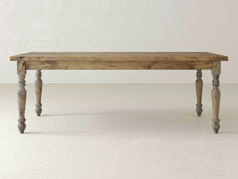 The French Country Dining Table - Harvest Wheat by James+James sits on a light-colored floor. The table showcases a rectangular top with a natural wood finish and has four intricately carved legs, giving it a classic, elegant appearance. The background features a plain, textured off-white wall.