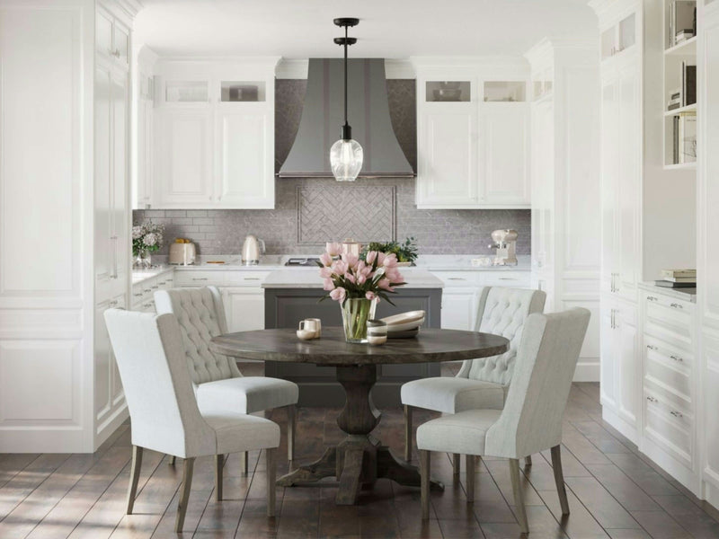 A modern kitchen features white cabinetry, a gray herringbone backsplash, and stainless steel appliances. In the center, a Sybil Round Dining Table - Deep Grey from James+James is surrounded by four cushioned chairs, and a vase of pink flowers decorates the table. The flooring is dark hardwood.