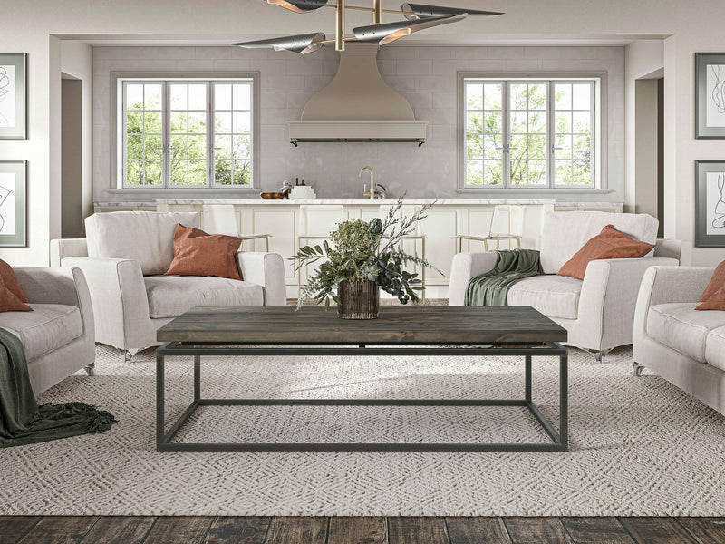 A modern living room features four light-colored armchairs with earthy-toned pillows and throws set around a Floating Top Coffee Table - Deep Grey by James+James on a textured rug. The room has two large windows letting in natural light, a minimalist kitchen in the background, and framed art on the walls.