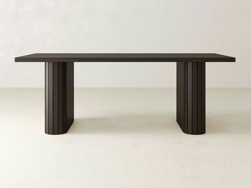 The Fern Dining Table - Charred Ember by James+James is a minimalist piece featuring a dark wooden, rectangular top supported by two solid, flat-panel legs. Its smooth surface rests elegantly on a light, plain background.