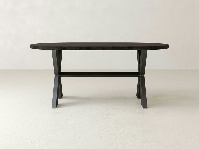 The X-Base Oval Dining Table - Charred Ember by James+James is a minimalist piece crafted from dark wood, featuring an oval top and sturdy, angular legs. Set against a plain, light-colored background, this table boasts a contemporary design with a crossbar for added stability and modern aesthetics.