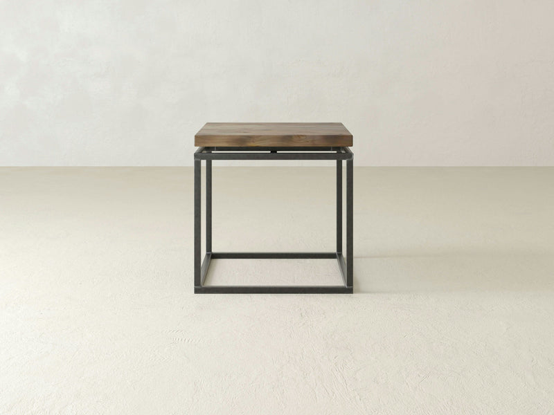 A minimalist end table named the Floating Top End Table - Barn Wood by James+James, featuring a wooden top and a black metal frame base, is positioned in an empty room with white walls and light-colored flooring. The design is simple and modern, emphasizing clean lines and functionality.