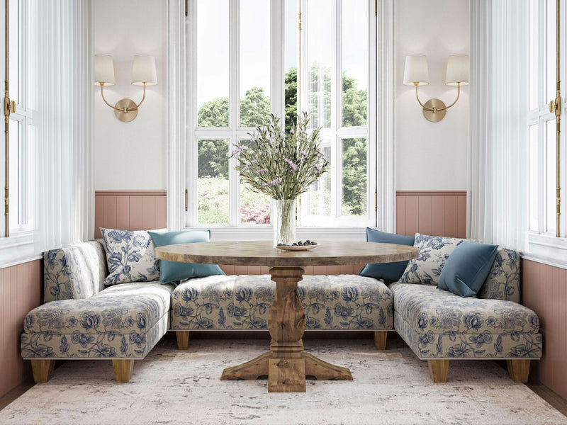 A cozy dining nook features the Heirloom Round Dining Table in Harvest Wheat from James+James, surrounded by U-shaped blue and white floral upholstered seating. The space is well-lit by large windows with white frames, and two wall sconces along with a view of lush green outdoor scenery contribute to the serene ambiance.