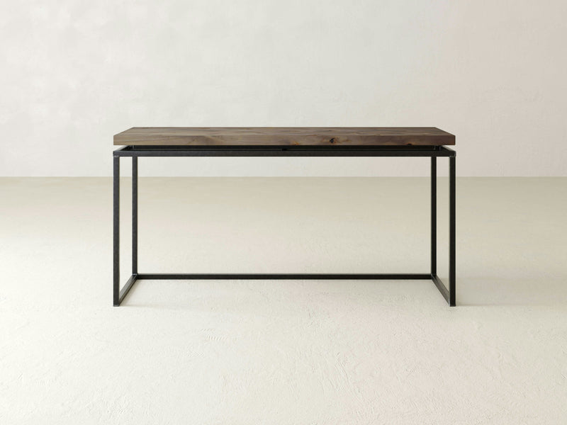 Against an off-white wall stands the Floating Top Sofa Table - Barn Wood by James+James. This minimalist console table features a dark brown top and a black metal frame, designed with clean lines and an open, rectangular structure for a simple and modern look.