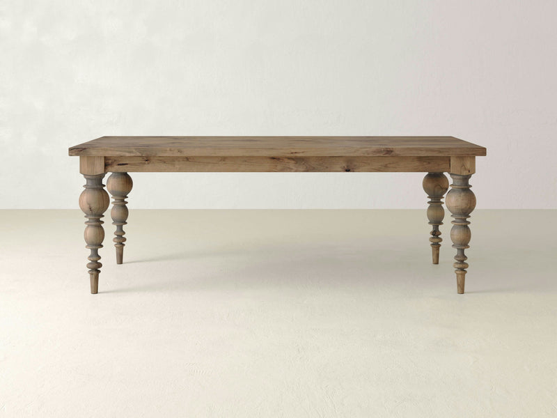 The James+James Bailey Dining Table boasts a rectangular wooden surface with a rustic finish and contemporary elegance. Featuring four modern turned legs, it stands on a light-colored floor against a plain, light gray wall. Its simple yet elegant design beautifully showcases the natural grain and texture of the wood.