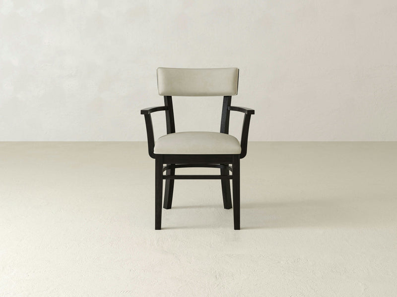 An Everly Arm Dining Chair - Charred Ember by James+James, featuring a black wooden frame and light-colored upholstery, is positioned in the center against a plain, light-colored background. The chair includes armrests and a slightly angled backrest designed for comfortable seating.