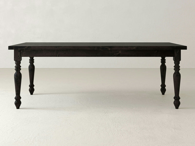 The French Country Dining Table - Charred Ember, crafted by James+James, features a rectangular dark wooden surface coupled with four ornately carved legs, standing elegantly against a plain, light-colored backdrop.