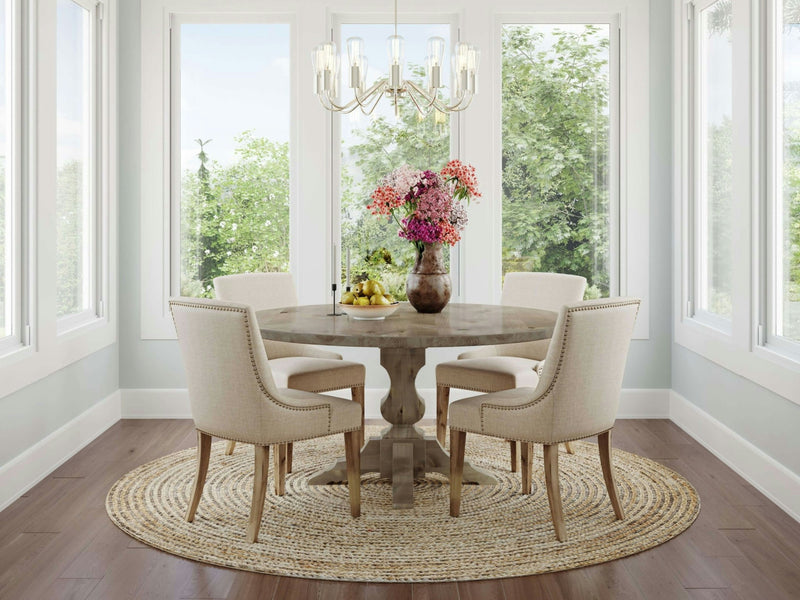 A light-filled dining room with large windows features the Sybil Round Dining Table - Barn Wood from James+James, centered on a woven rug. The table is surrounded by four cushioned chairs and adorned with a vase of colorful flowers and a bowl of fruit. A modern chandelier hangs overhead.