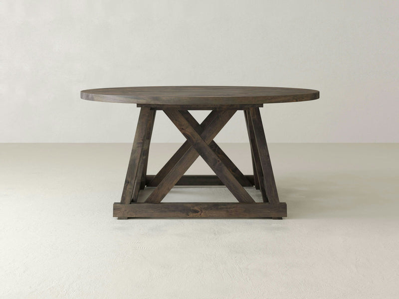 The Julia Round Dining Table - Deep Grey by James+James stands on a unique cross-braced base. The design features a simple, rustic style with a sturdy and solid structure, perfect for dining or as a centerpiece in a living space. The background is a plain, light-colored wall and floor.