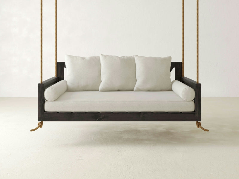 The Avery Porch Swing - Charred Ember by James+James is a minimalist hanging swing bed with a dark wooden frame, suspended by thick ropes. It features cream-colored seat cushions, three large back pillows, and two cylindrical side pillows, set against a plain white wall.