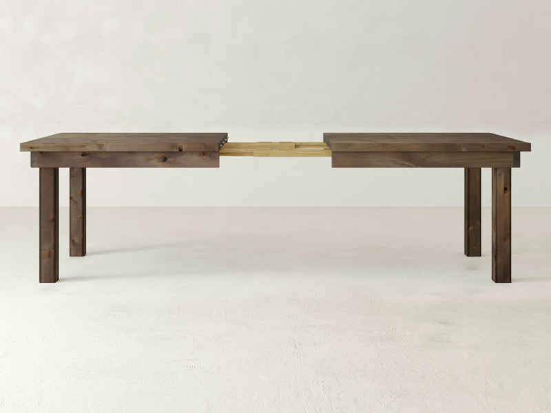 Introducing the Farmhouse Expandable Dining Table - Barn Wood by James+James. This table features a rustic finish, four sturdy legs, and a rectangular shape. It boasts a visible central extension mechanism designed to accommodate additional leaves for added length. The backdrop includes a plain beige wall and floor.