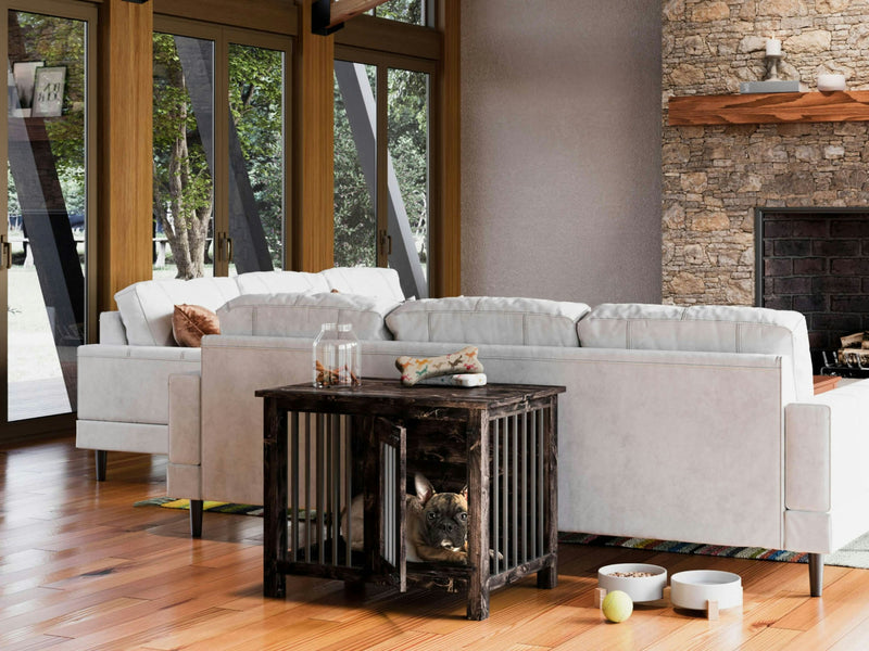 A modern living room features a large white sectional sofa facing a stone fireplace. A dog rests in the Single Dog Kennel - Charred Ember by James+James, which doubles as a side table next to the sofa. The room has large windows with views of outdoor greenery, and toys are scattered on the hardwood floor.