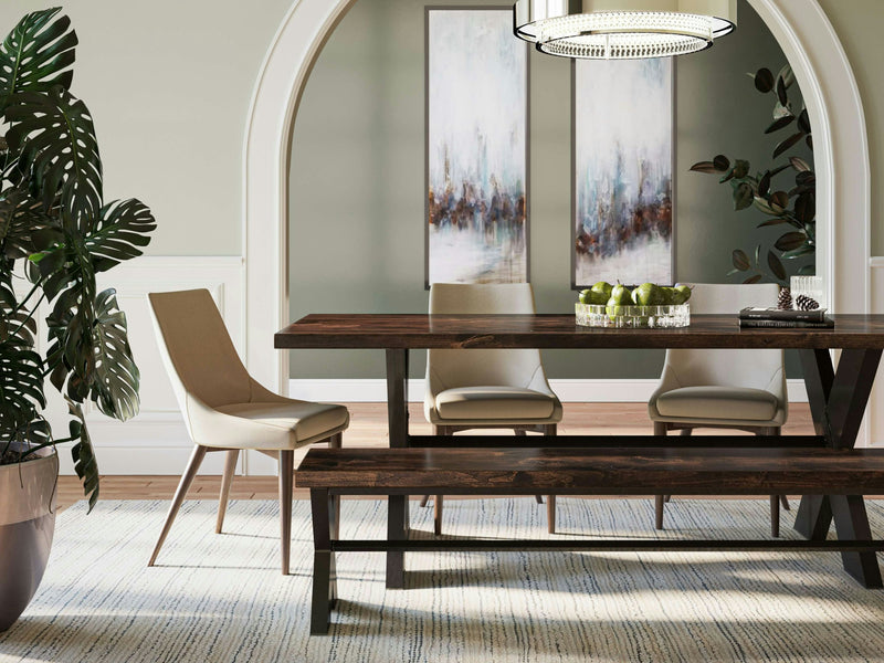 A modern dining room featuring a stylish dark wooden table paired with the X-Base Bench in Tobacco from James+James, along with two beige chairs. The table is adorned with a bowl of green apples, while the light green walls are decorated with two abstract paintings. A large potted plant sits to the left, and an arched doorway can be seen in the background.