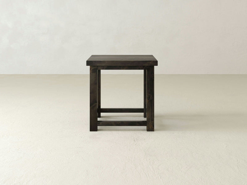 The Carter X Side Table in Deep Grey from James+James is a minimalist piece with a flat top and four legs connected by horizontal supports. The table stands alone in a plain, light-colored room with a smooth floor and a simple, unadorned background.