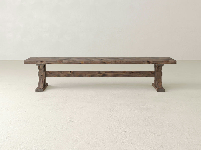 The Trestle Bench - Barn Wood by James+James features a simple, rustic design with a natural finish. It boasts a flat, rectangular seat supported by two sturdy legs at each end, connected by a horizontal brace. The bench is set against a plain, off-white background.