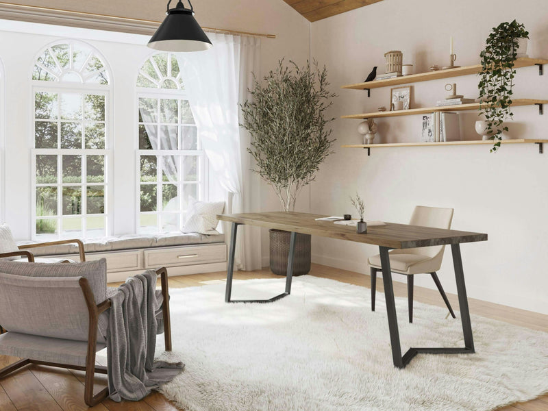 A bright, modern home office with large arched windows features the Arkwright Desk in Harvest Wheat from James+James, complemented by a white rug. A potted plant stands beside the desk. Shelves adorned with plants and decor items are mounted on the wall. A cozy reading nook with a window seat and a cushioned chair is visible.