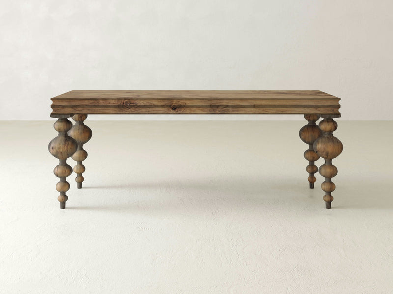 The Olivia Sofa Table - Harvest Wheat by James+James is a wooden table with a rectangular top showcasing a natural wood grain finish. It's supported by four uniquely designed legs, each carved into a combination of spherical and cylindrical shapes, creating an artistic and elegant look against a plain, off-white background.