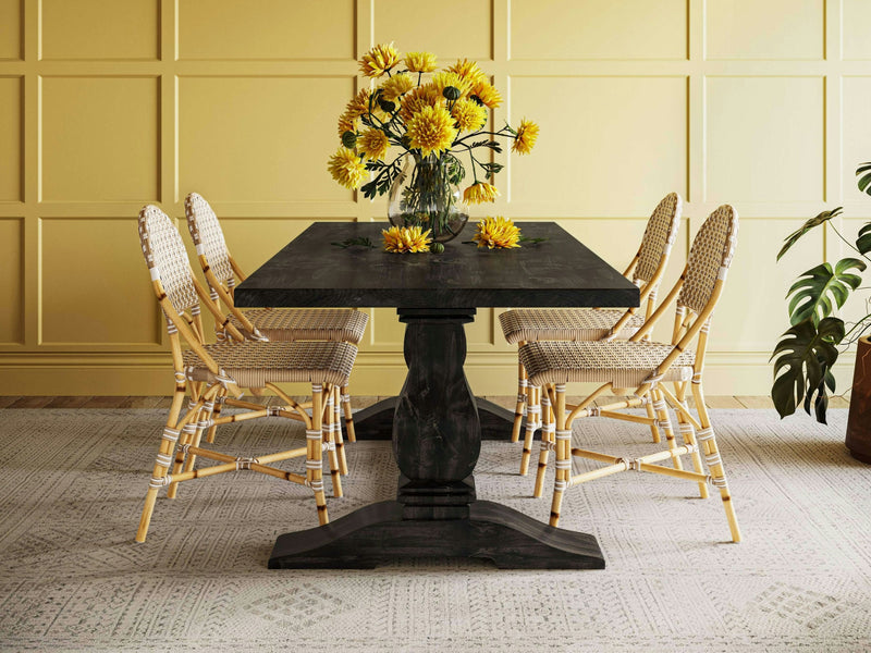 A dining room showcasing the Heirloom Dining Table - Charred Ember by James+James is surrounded by six woven wicker chairs. The table is adorned with a vase filled with yellow flowers, and a few loose flowers are artistically scattered on its surface. A yellow paneled wall and a potted plant provide a charming backdrop.