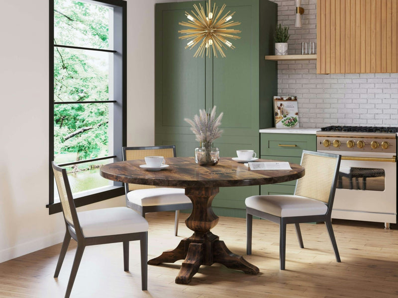 A cozy dining area features the Sybil Round Dining Table in Tobacco by James+James, surrounded by four light-colored chairs. A modern sputnik-style chandelier hangs above. The backdrop includes green cabinets, a white brick backsplash, and large windows offering a view of lush greenery outside.