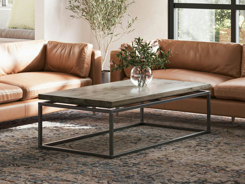 A living room with two brown leather sofas facing each other and a James+James Floating Top Coffee Table in Barn Wood in between. A glass vase with flowers is on the table. Sunlight filters through large windows, illuminating the room and an ornate rug underneath.