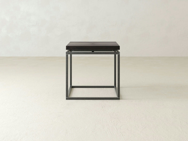 The Floating Top End Table - Charred Ember by James+James is a minimalist black metal side table featuring a square frame and a wooden top, set against a plain, light-colored background.