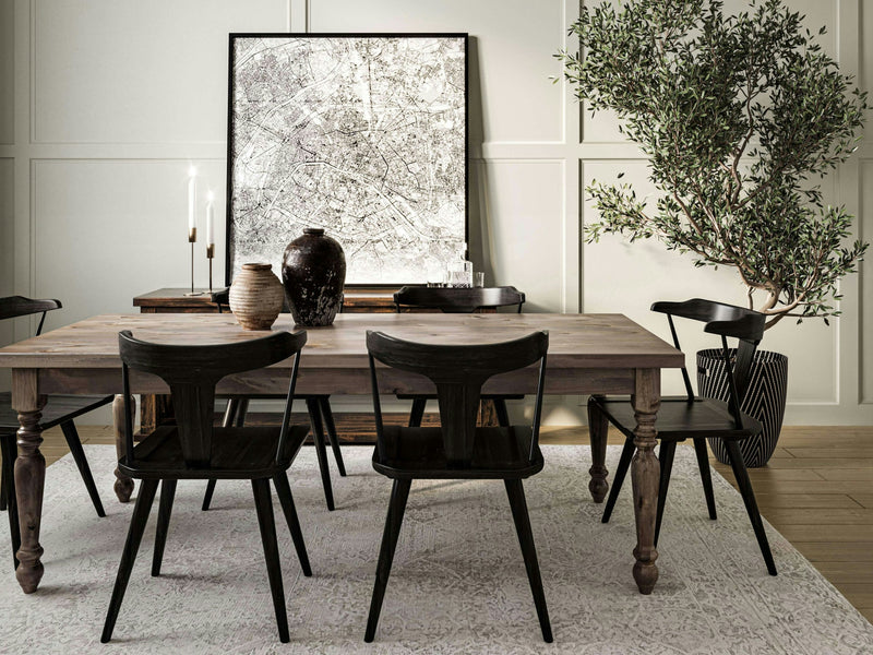 A dining area features the French Country Dining Table - Barn Wood from James+James accompanied by four black chairs. On the table are two candles, a dark vase, a brown jug, and an abstract artwork. An ornamental tree in a planter stands beside the table, all set against a paneled wall in a light-colored room.