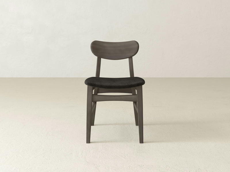 The Lynn Upholstered Dining Chair - Deep Grey by James+James is centered in the image. This modern, minimalist wooden chair features a curved backrest and sleek, dark finish. It has a black cushioned seat and stands on light, neutral-colored flooring against a plain, off-white wall.