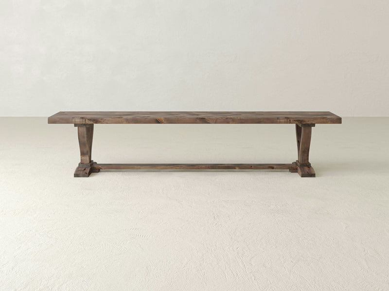 The Vera Bench - Barn Wood by James+James is a rustic wooden bench featuring a rectangular seat and V-shaped legs supported by a horizontal beam. The weathered wood gives it a vintage look, and its simple design showcases a natural, minimalist aesthetic.