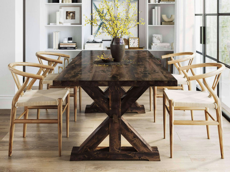 A dining area featuring the Trestle Dining Table - Tobacco by James+James with six wicker chairs arranged around it. The table is adorned with a dark brown vase holding yellow floral branches. In the background, a white bookshelf displays various decor items, and a large window allows natural light to pour in.