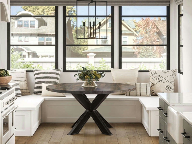 A cozy kitchen nook featuring the Shiloh Round Dining Table in deep grey by James+James, with a stylish black geometric base, surrounded by white built-in bench seating adorned with patterned pillows. The space is illuminated by large windows showcasing a suburban neighborhood and modern pendant lighting above.