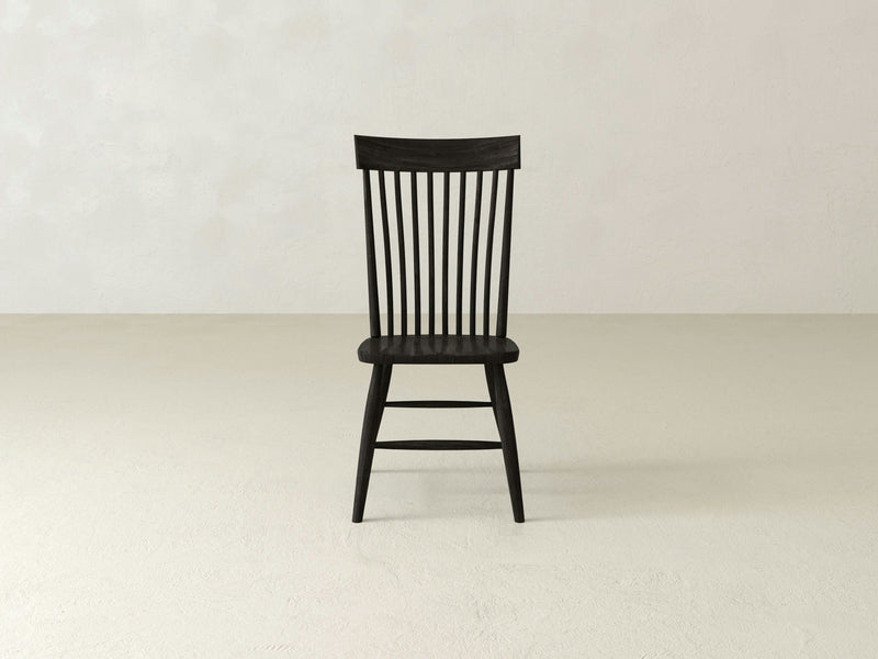 A solid wood Eleanor Side Dining Chair in black, crafted by James+James, stands on a plain, light-colored floor against a light gray wall. The chair features vertical slats on the backrest and four straight legs, embodying a simple and minimalist design that exudes a timeless aesthetic.