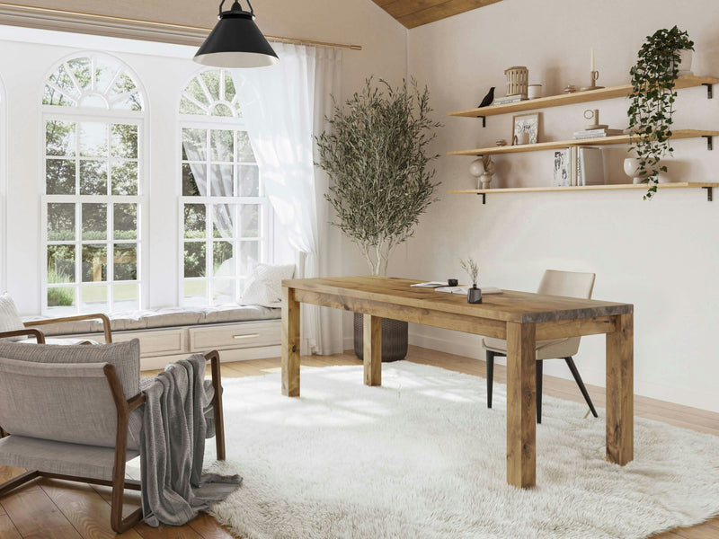 A cozy home office featuring the Jonathan Desk in Harvest Wheat by James+James, a beige chair, and a cream rug on hardwood floors. There's a window seat with cushions and a tall indoor plant beside the desk. Shelves with decor items and a hanging light are on the right wall, while light pours in through arched windows.