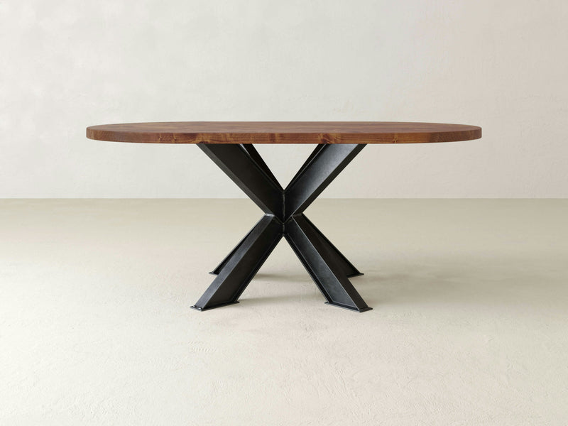 The Shiloh Oval Dining Table - Tuscany by James+James features a dark brown wooden top and black metal geometric legs that intersect to form a durable, contemporary base. This table is set in a minimalist, light-colored room with no other furniture or decorations visible.