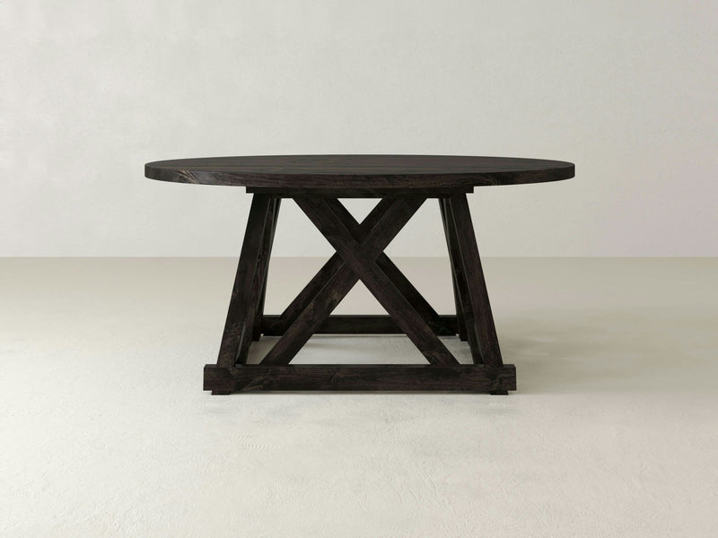 Against a plain white background, the Julia Round Dining Table - Charred Ember by James+James stands with its sturdy X-shaped base. Made of dark wood, this table boasts a simple yet rustic design and rests on a light-colored floor.