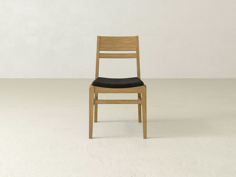 Introducing the Delilah Dining Chair - Harvest Wheat by James+James: a wooden chair with a minimalistic design, featuring a black cushioned seat and a backrest with two horizontal slats. This elegant chair is showcased against a plain, light-colored background.