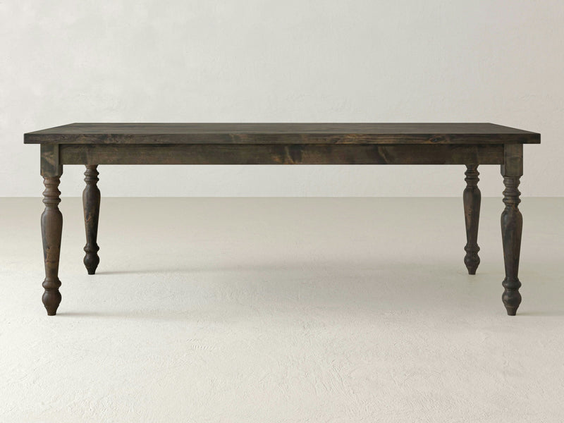 A French Country Dining Table - Deep Grey by James+James, featuring a rectangular top and turned legs, stands on a light-colored, plain floor with a light-colored wall in the background. The table's design exudes a rustic and traditional appearance.