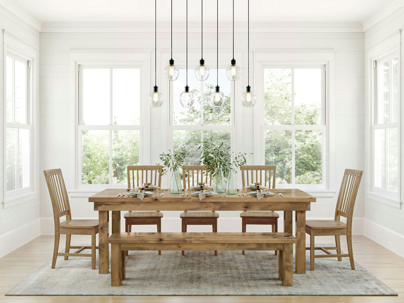 A dining room showcases the Farmhouse Expandable Dining Table - Harvest Wheat from James+James, complete with four matching chairs and a bench. The table is beautifully decorated with tableware and vases filled with greenery. Seven pendant lights dangle above, casting a warm glow, while large windows flood the room with natural light and offer picturesque views of trees outside. The space exudes a light, airy ambiance.