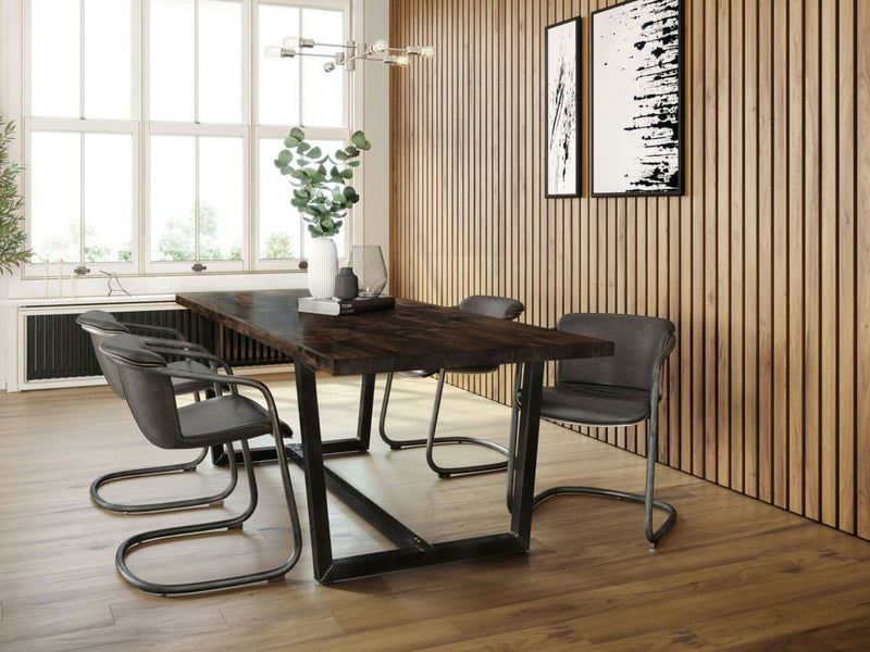 A modern dining room featuring the Trapezoid Dining Table - Tobacco by James+James, accompanied by four sophisticated gray chairs. The table showcases a dark brown top and sturdy metal legs. The walls boast vertical wooden paneling, complemented by two abstract black-and-white paintings on the right wall. A large window bathes the space in natural light.